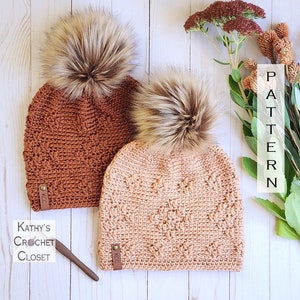 May include: Two crocheted hats with faux fur pom poms. The hats are in shades of brown and peach. The hats have a label that says "KCC" on them. The hats are on a white wooden surface. The text "PATTERN" is visible in the background.