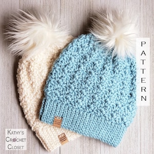 May include: Two crocheted hats with faux fur pom poms. The hats are in a light blue and a cream color. The hats have a label that says "Kathy's Crochet Closet".