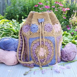 K&ouml;nnte beinhalten: Handgefertigter H&auml;kelrucksack mit Kordelzug in Beige, Blau und Lila. Die Tasche hat Blumenmuster und einen Kordelzugverschluss. Kugeln aus rosa und blauer Wolle umgeben die Tasche, mit einem Hintergrund aus Blumen und Gr&uuml;n.