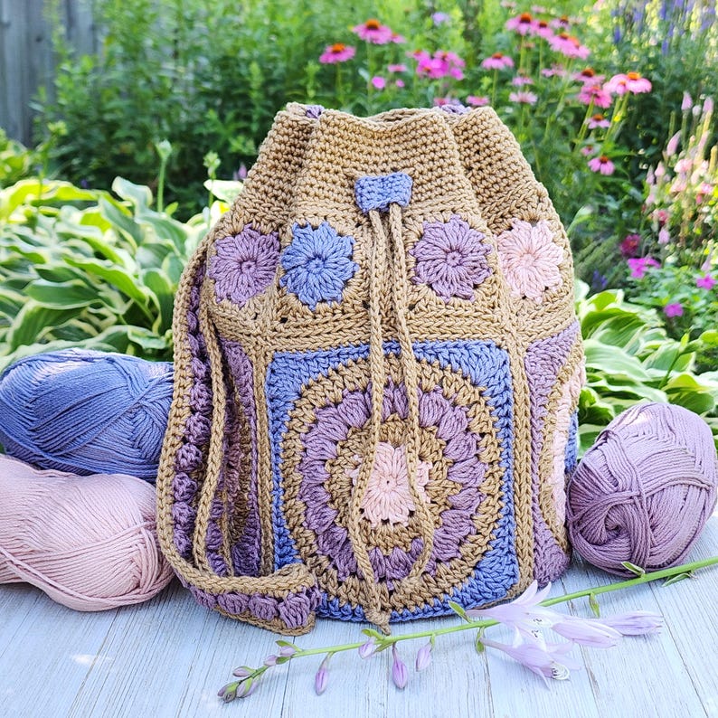 K&ouml;nnte beinhalten: Handgefertigter H&auml;kelrucksack mit Kordelzug in Beige, Blau und Lila. Die Tasche hat Blumenmuster und einen Kordelzugverschluss. Kugeln aus rosa und blauer Wolle umgeben die Tasche, mit einem Hintergrund aus Blumen und Gr&uuml;n.