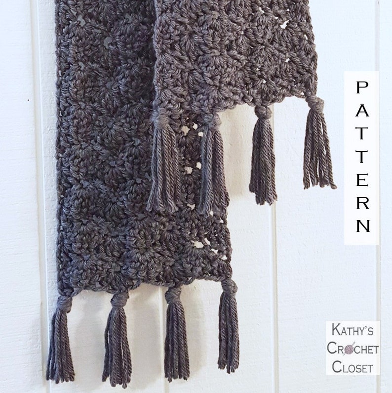 May include: A gray crocheted scarf with tassels on the ends. The scarf is hanging on a white wall. The text "PATTERN" is written vertically on the wall.