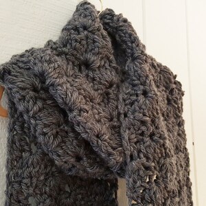 May include: A gray crocheted scarf with a textured pattern. The scarf is hanging on a wooden hanger.