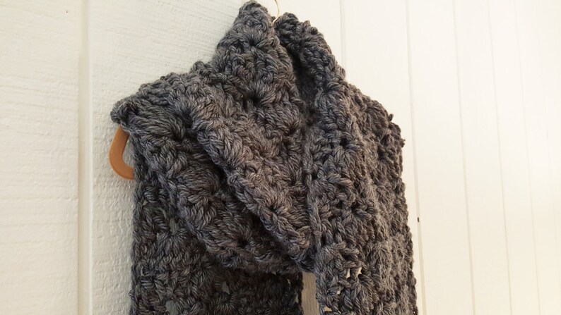 May include: A gray crocheted scarf with a textured pattern. The scarf is hanging on a wooden hanger.