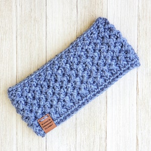Op de afbeelding: Een blauwe gehaakte hoofdband met een bruin leren label waarop "Kathy's Crochet Closet" staat.