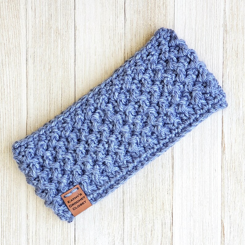 Op de afbeelding: Een blauwe gehaakte hoofdband met een bruin leren label waarop "Kathy's Crochet Closet" staat.
