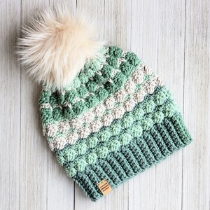 May include: A green, white and light green crocheted beanie with a fluffy white pom pom on top. The beanie has a small brown tag attached to it that says "Katie's Crochet Closet".