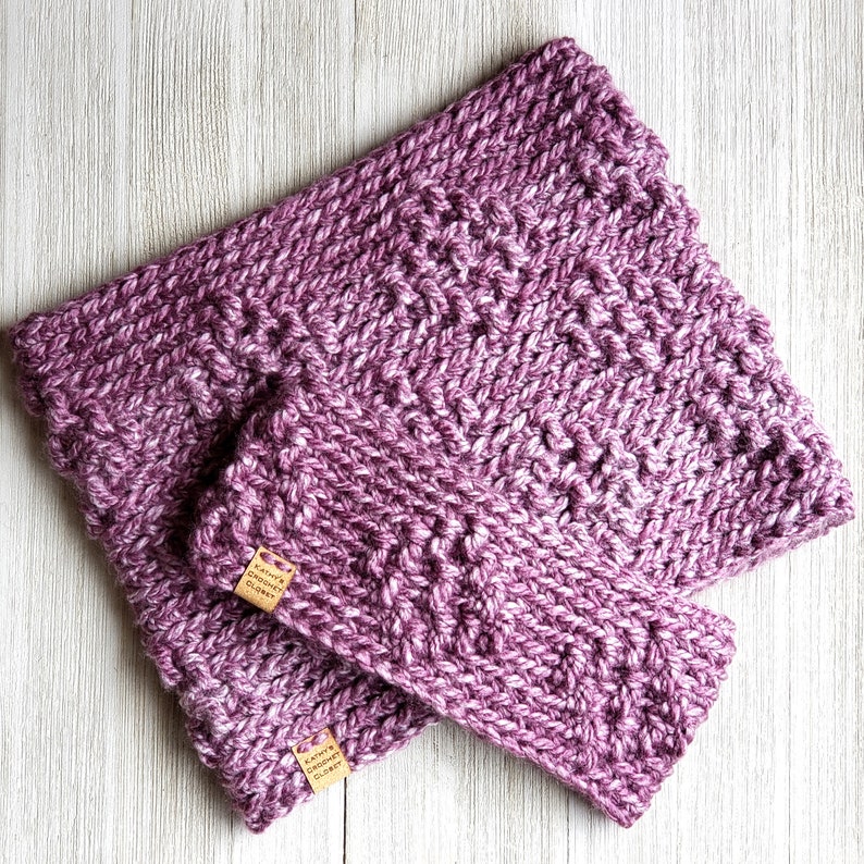 Op de afbeelding: Een paarse gebreide sjaal en bijpassende vingerloze handschoenen met een bruin lederen label met de tekst "Kathy's Knits Closet".