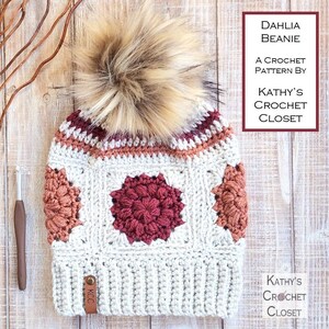 May include: A crocheted beanie hat with a faux fur pom pom in cream, burgundy, and peach colors. The beanie has a square pattern with a large burgundy flower in the center. The hat has a leather tag that says "KCC". The text "DAHLIA BEANIE A CROCHET PATTERN BY KATHY'S CROCHET CLOSET" is visible in the upper right corner of the image.