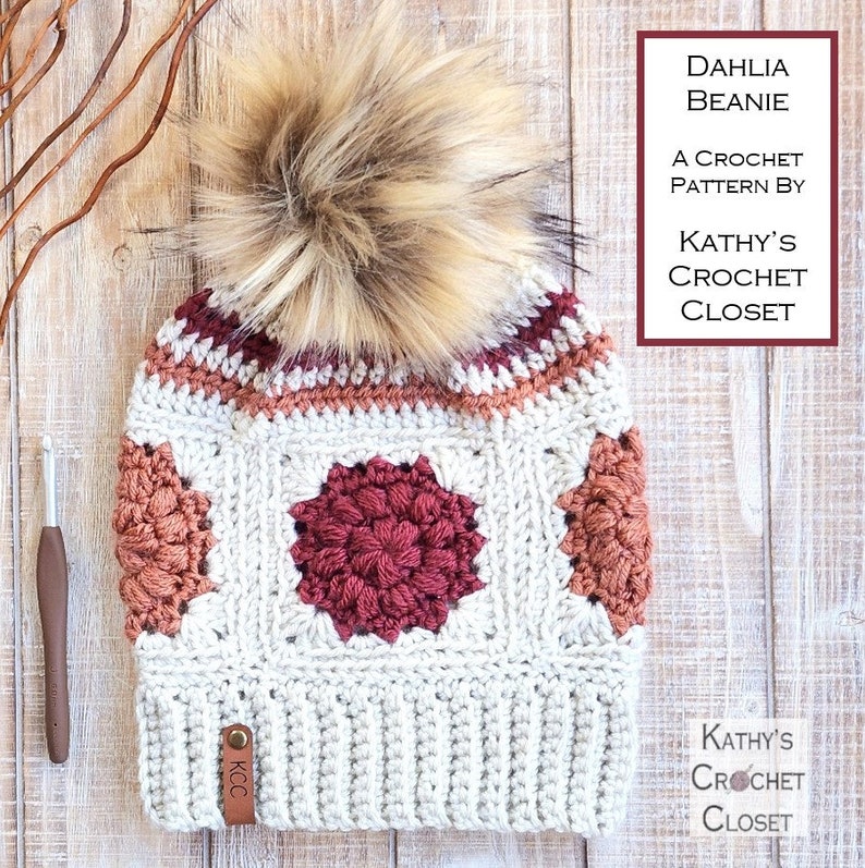May include: A crocheted beanie hat with a faux fur pom pom in cream, burgundy, and peach colors. The beanie has a square pattern with a large burgundy flower in the center. The hat has a leather tag that says "KCC". The text "DAHLIA BEANIE A CROCHET PATTERN BY KATHY'S CROCHET CLOSET" is visible in the upper right corner of the image.