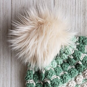 May include: A green and white crocheted hat with a fluffy cream-coloured pom pom on top.