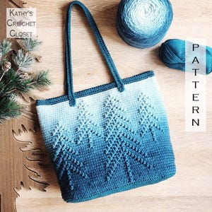 May include: A crochet tote bag featuring an ombre blue color gradient and a textured pine tree pattern. The bag has two shoulder straps and is displayed with two balls of blue yarn and a wooden tree cutout. Text reads Kathy's Crochet Closet.