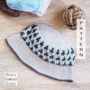 May include: A crocheted bucket hat in light gray with a geometric pattern in blue and brown. The hat has a blue and brown trim. The text "PATTERN" is visible. The text "KATHY'S CROCHET CLOSET" is also visible.