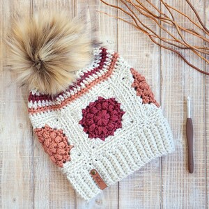 May include: A crocheted beanie hat with a white base and colorful geometric patterns in shades of pink, red, and orange. The hat has a fluffy brown pom pom on top and a brown leather tag with the letters "KCC" on the side.