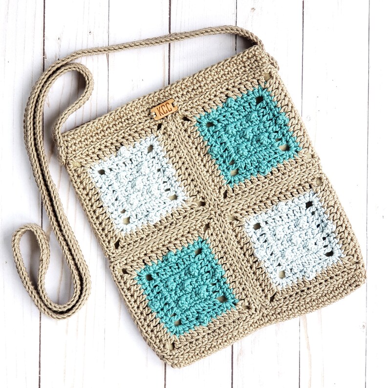 Puede incluir: Un bolso de hombro tejido a crochet con una correa marr&oacute;n y un dise&ntilde;o de granny squares marr&oacute;n, blanco y turquesa. El bolso tiene una peque&ntilde;a etiqueta de cuero con las letras "KCC" en ella.
