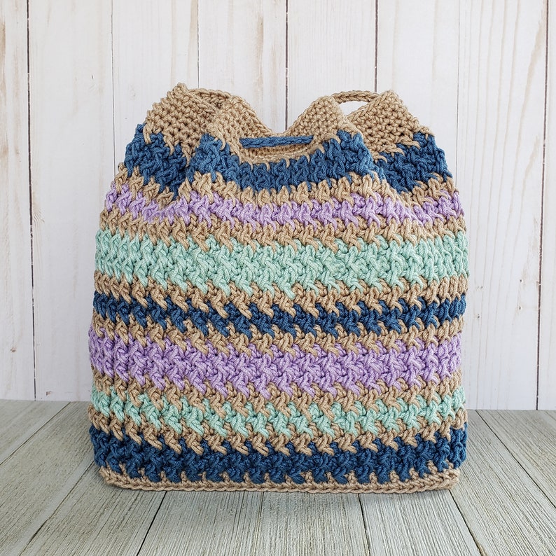 Pu&ograve; includere: Una borsa a mano all'uncinetto con un motivo a righe in tonalit&agrave; di blu, verde, viola e marrone. La borsa &egrave; realizzata con un filato testurizzato e ha una chiusura con coulisse.
