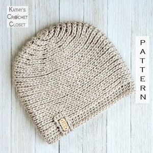May include: A beige crocheted beanie with a small wooden tag that says "KCP" on it. The beanie is on a white wooden surface. PATTERN