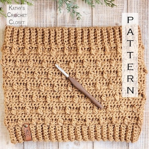 May include: A close-up of a crocheted hat in a brown color with a leather tag that says "KCC". The hat is lying on a wooden surface with a crochet hook next to it. The text "PATTERN" is written vertically on the right side of the image.