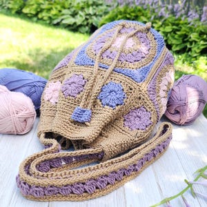 K&ouml;nnte beinhalten: Handgefertigter H&auml;kelrucksack in Beige, Lavendel und Blau. Die Tasche hat einen Kordelzugverschluss, einen langen Riemen und ist mit Blumenmotiven verziert. Mehrere Kn&auml;uel Garn in Rosa, Lavendel und Blau sind ebenfalls zu sehen.
