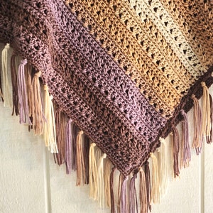 Puede incluir: Un chal triangular de ganchillo con un patr&oacute;n de colores degradados que transita de marr&oacute;n oscuro a malva, beige y crema. El tejido presenta un dise&ntilde;o de puntos calados y est&aacute; rematado con largos flecos anudados a juego.