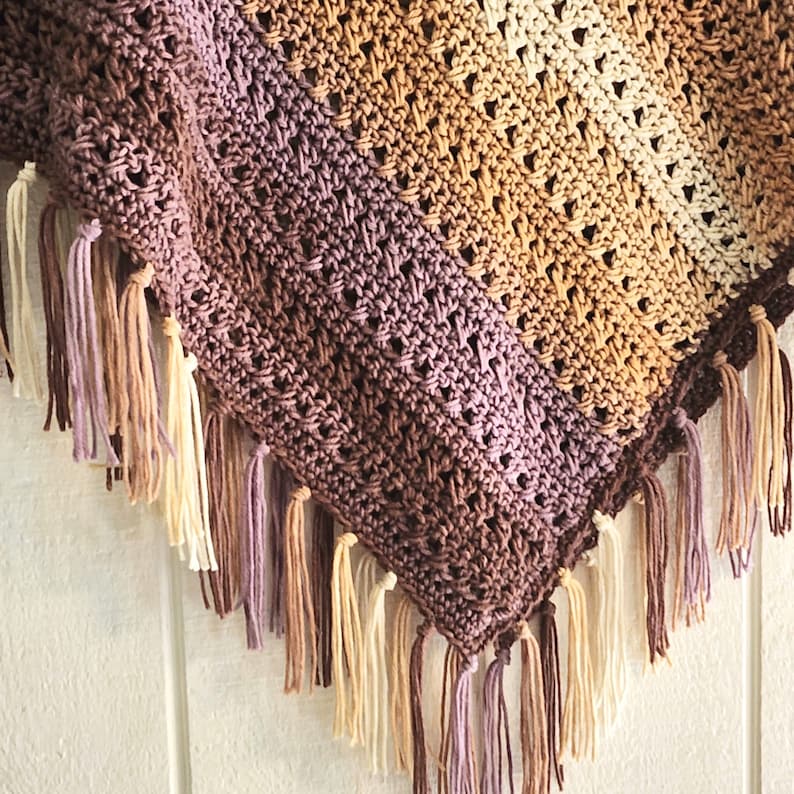 Puede incluir: Un chal triangular de ganchillo con un patr&oacute;n de colores degradados que transita de marr&oacute;n oscuro a malva, beige y crema. El tejido presenta un dise&ntilde;o de puntos calados y est&aacute; rematado con largos flecos anudados a juego.