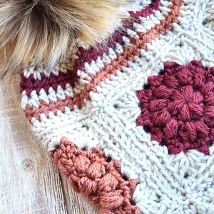 May include: A crocheted hat with a pompom made of faux fur. The hat is made with a variety of colors, including white, beige, brown, and red.