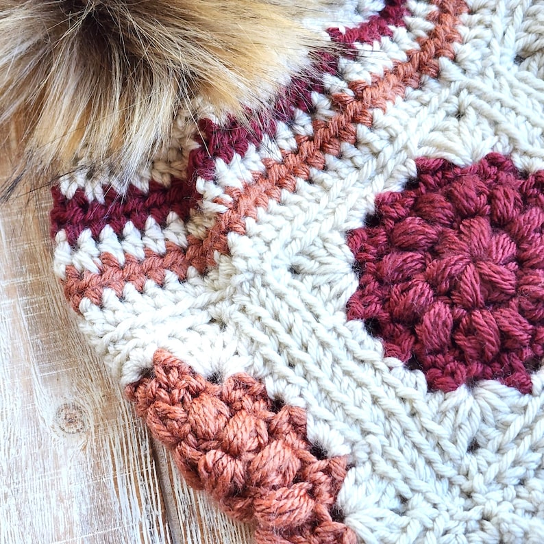 May include: A crocheted hat with a pompom made of faux fur. The hat is made with a variety of colors, including white, beige, brown, and red.
