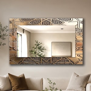 Op de afbeelding: Een rechthoekige wandspiegel met een brede, reflecterende lijst met een herhalend geometrisch patroon in brons- en donkerbruintinten. Het spiegeloppervlak reflecteert een lichte binnenruimte met een raam, een plant en een ingelijst kunstwerk.