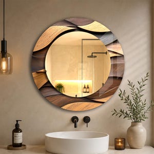 May include: A round wall mirror featuring a decorative frame with layered, wavy wooden panels in shades of brown and tan. The mirror reflects a bathroom interior with a white vessel sink, a faucet, and a light-coloured countertop.