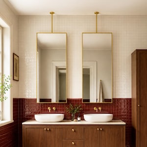 Op de afbeelding: Een dubbel badkamermeubel met een donkere houten afwerking, twee witte ovale waskommen en goudkleurige kranen. Boven het meubel hangen twee rechthoekige spiegels met gouden frames aan het plafond. Een hoge bijpassende houten kast staat ernaast.