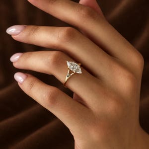 Op de afbeelding: Een gouden ring met een enkele transparante edelsteen in marquise-slijpvorm in een klauwzetting. De ringband heeft een gespleten ontwerp dat samenkomt bij de basis van de steen, met een gepolijste geelgouden afwerking en een gefacetteerde steen.