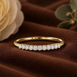 Könnte beinhalten: Ein dünner Goldring mit einer Reihe von zehn kleinen, runden, klaren und facettierten Steinen, die entlang der oberen Hälfte des Rings eingefasst sind. Das Metall hat eine polierte, reflektierende Oberfläche und die Steine sind gerade.