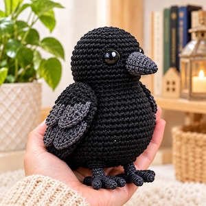 Könnte beinhalten: Eine kleine, handgefertigte Häkelfigur in Form eines Vogels aus schwarzem Garn. Der Vogel hat einen runden Körper, einen strukturierten grauen Schnabel und dunkelgraue Flügeldetails. Er besitzt große, glänzende schwarze Sicherheitsaugen und kleine Füße.