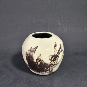 May include: A small, round, off-white ceramic vase with a smooth, glossy finish. The exterior features a dark brown, hand-painted illustration of a bird with spread wings and detailed feather patterns. The interior of the vase is dark brown.