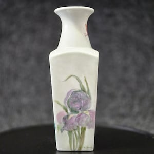 May include: A white ceramic vase with a square base and a flared neck. The front of the vase features a hand-painted illustration of a purple iris flower with green stems and leaves. The surface has a smooth finish with a subtle floral design.