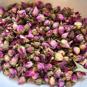 May include: A collection of dried rose buds in shades of pink, magenta, and cream. The buds are clustered together in a white container. Text printed above the rose buds reads: Available from 5 g to 500 g. The dried flowers have green sepals.