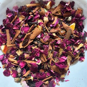 Può includere: Una miscela di tè sfuso con petali di rosa magenta essiccati, bastoncini di corteccia di cannella marroni, chiodi di garofano marrone scuro e piccoli pezzi di scorza d'arancia essiccata. Il testo "Available from 5 g to 500 g" è stampato in rosso scuro.