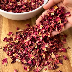May include: A collection of dried rose petals in shades of deep magenta, crimson, and pale yellow. The petals are scattered across a wooden surface, with a portion contained in a white bowl and a handful held in an open palm.