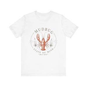 Può includere: Una t-shirt bianca a maniche corte con una grafica circolare sul petto. Il design include un'illustrazione di un gambero di fiume color bruno-rossastro, canne e il testo MUDBUG STATE OF MIND con EST. e LA ai lati dell'emblema circolare centrale.