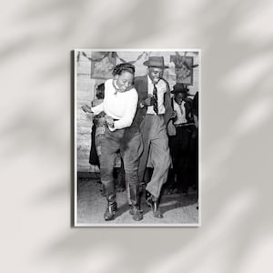 Puede incluir: Una fotografía en blanco y negro que muestra a dos personas bailando en una habitación. Una persona viste una blusa de manga larga clara, pantalones oscuros y botas, mientras que la otra lleva un traje oscuro, camisa, corbata y un sombrero fedora.