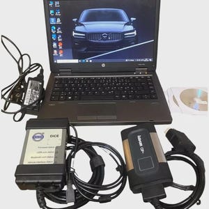 May include: A dark grey laptop with a Volvo car image on the screen, accompanied by a black power adapter, two diagnostic interface units labelled Volvo DiCE and Auto-com CDP+, associated black connection cables, and two optical discs.