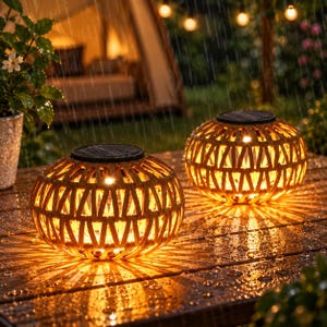 May include: Two round solar lanterns with a woven tan exterior and a dark top panel. The lanterns emit a warm, glowing light through the lattice pattern, casting rays onto a wet wooden surface during a rain shower in an outdoor setting.