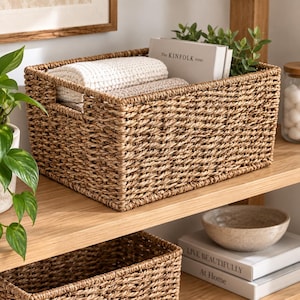 Puede incluir: Una cesta de almacenamiento rectangular de algas marinas tejidas con asas laterales integradas. La cesta contiene un tejido de color crema con textura doblado, un libro titulado The KIN F O L K HOME y una pequeña planta verde de hojas ovaladas.