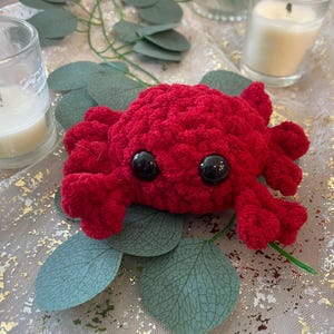 May include: A small, handmade crochet crab crafted from vibrant red yarn. The plush toy features a rounded body with multiple textured legs extending from the sides and two large, smooth, black plastic safety eyes positioned on the front.