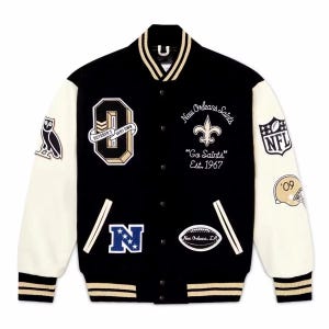 Op de afbeelding: Een zwart varsity jack met crèmekleurige mouwen, voorzien van geborduurde patches waaronder een uil, de letter O met de tekst October's Very Own, het New Orleans Saints logo, het NFL-schild, een football en de tekst Go Saints Est. 1967.