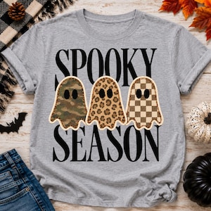 Może przedstawiać: Szary melanżowy t-shirt z czarnym napisem SPOOKY SEASON. Pomiędzy słowami znajdują się trzy kształty duchów z czarnymi owalnymi oczami. Pierwszy duch ma zielony wzór moro, drugi brązowy wzór w panterkę, a trzeci brązowo-beżowy wzór w szachownicę.