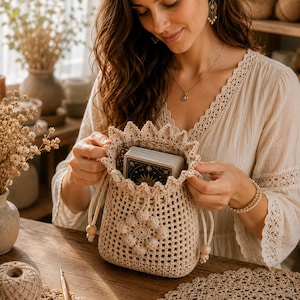 Puede incluir: Una bolsa de ganchillo beige con cordón, borde superior festoneado y un motivo floral central. La bolsa está construida con un patrón de rejilla abierta y contiene una baraja de cartas del tarot con ilustraciones celestiales azul oscuro y oro.