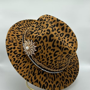 Puede incluir: Un sombrero fedora con estampado de leopardo en marrón y negro. El sombrero está adornado con una cinta negra, un cordón trenzado en tono dorado, una hilera de pequeñas cuentas blancas y un gran broche en forma de estrella con cristales.
