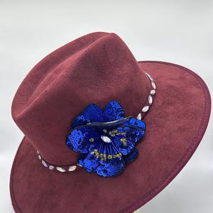 May include: A deep burgundy fedora hat with a matte finish, featuring a large, vibrant blue sequined flower embellishment on the front. The hat band is decorated with a series of clear, faceted crystals that catch the light along the brim.