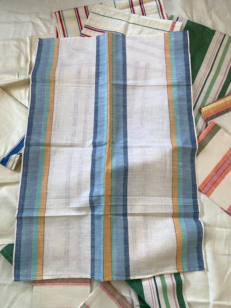 May include: A collection of white linen towels with vertical stripes in various colors. The central towel features stripes of blue, teal, and orange. Other towels display stripes of red, green, and pink. The towels are folded and arranged on a white surface.