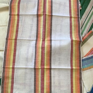 May include: White linen towel with vertical stripes in shades of orange, yellow, and brown. Other striped towels in various colors are visible in the background. The towel is folded and appears to be new.